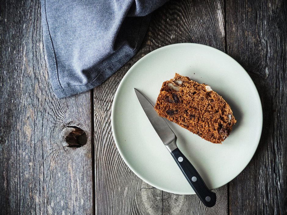 A slice of rosemary bread on a white plate with a knife on a wood surface with a blue linen.