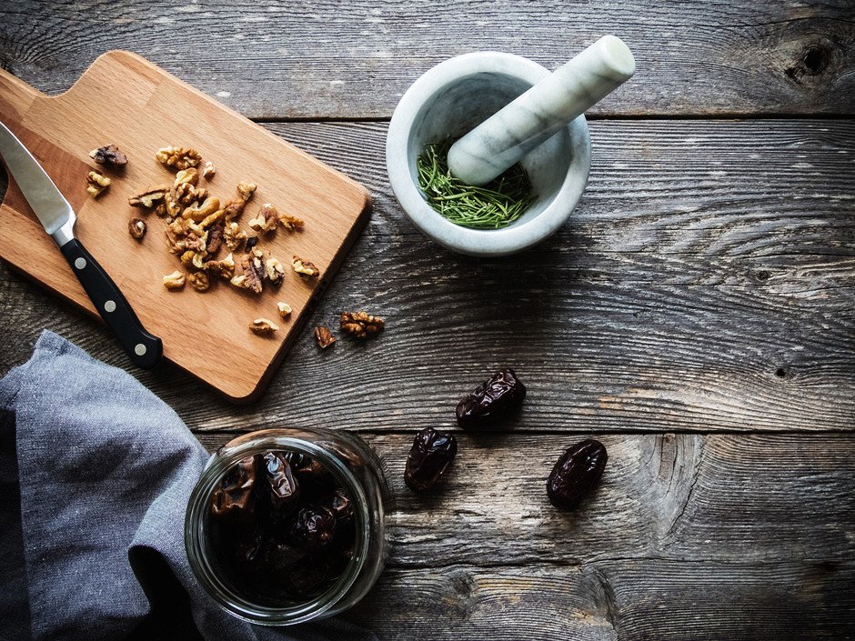 walnuts on a cutting board, a mortar and pestle with rosemary and a jar of dates on a wood surface.