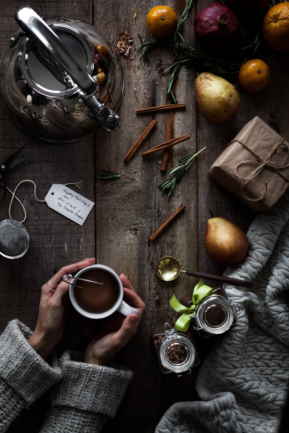 overhead of two hands cupping a mug of tea with a kittle, spices, fruit and tea jars on a wood surface.