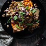 overhead of a stuffed sweet potato on a black plate with a linen and bowl of dressing with a spoon.