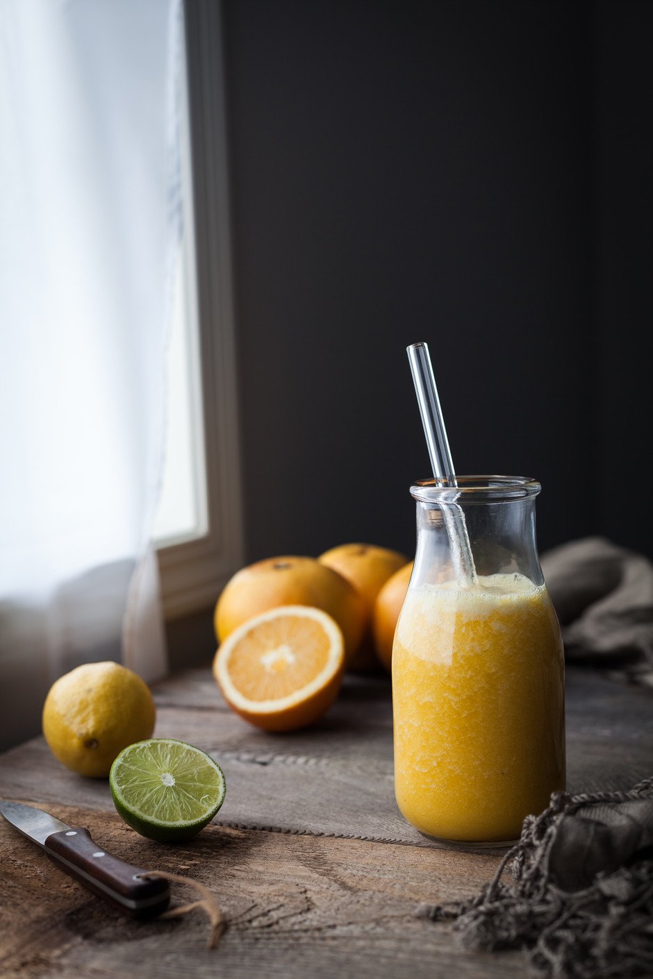 a glass of orange juice with a straw next to window with citrus.