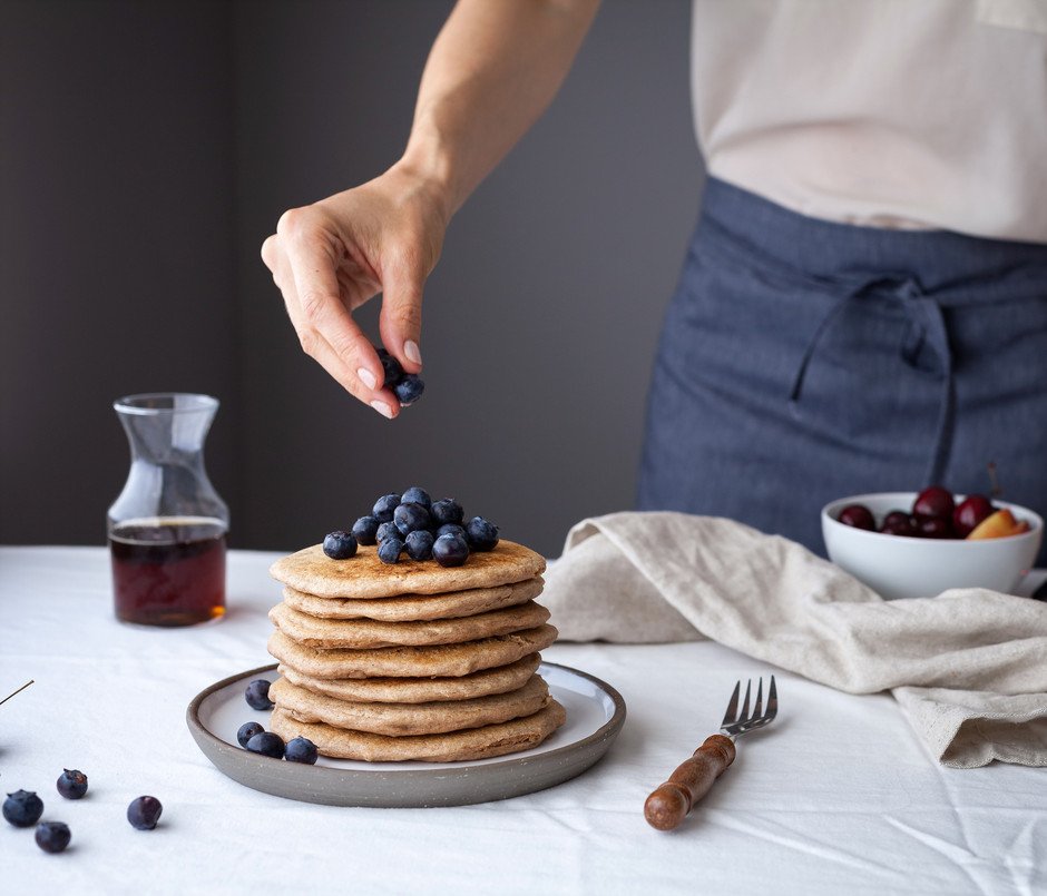 a baker placing blueberries on top of a stack of fluffy vegan spelt pancakes.