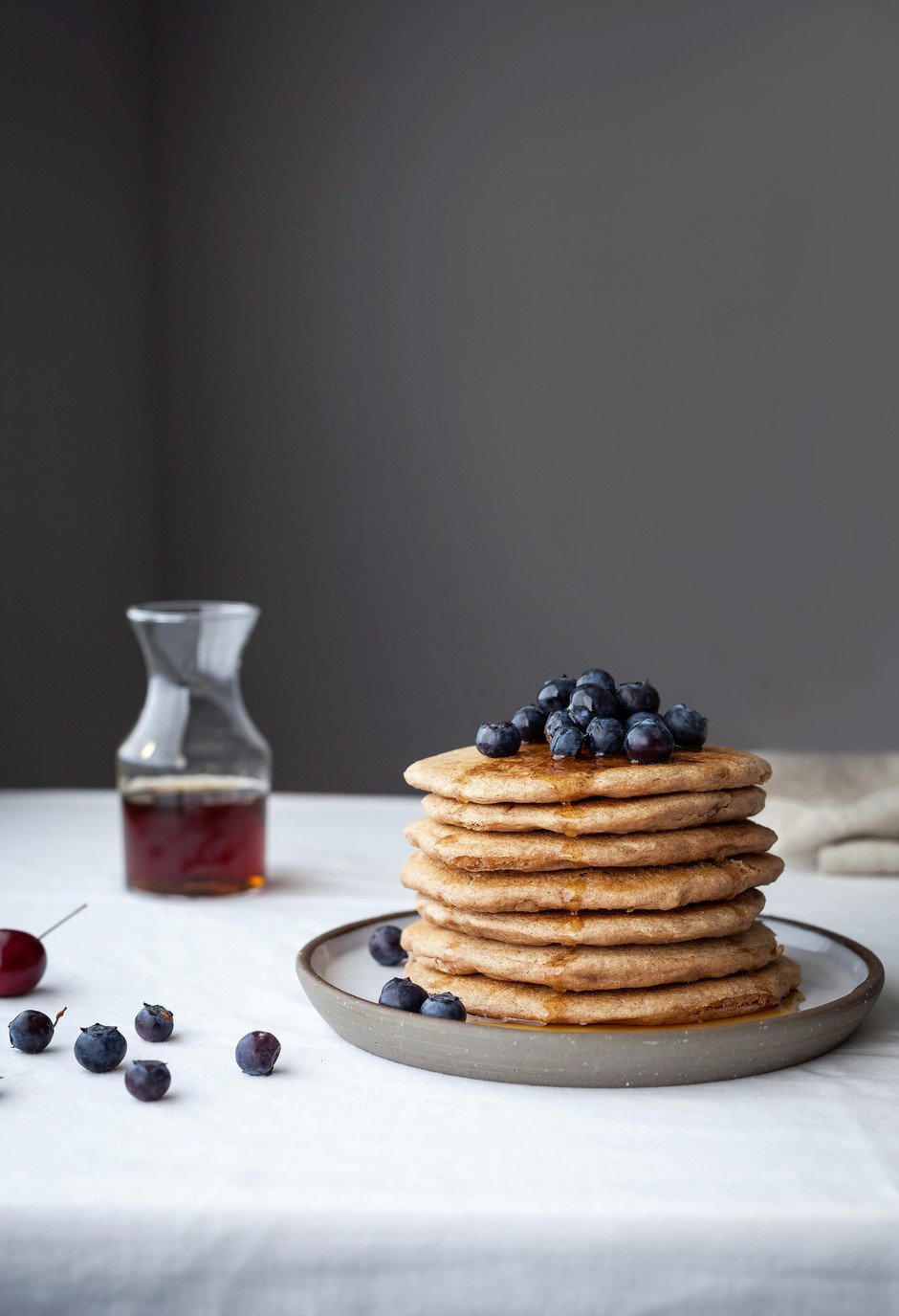 a close up of a stack of vegan spelt pancakes with blueberries and maple syrup on top.