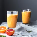 two glasses of orange smoothie with cut citrus fruits on a marble surface with a linen.