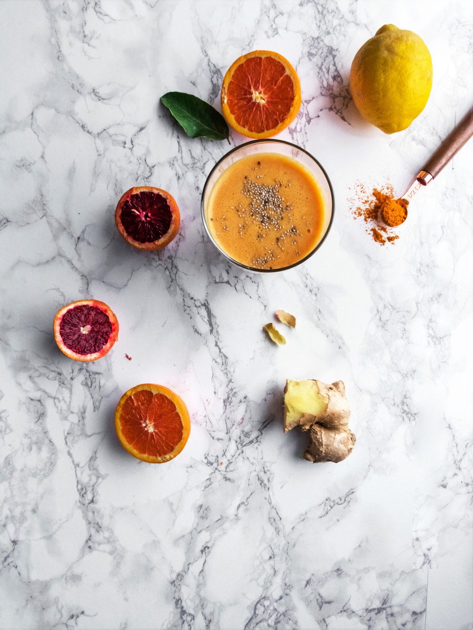 Overhead image of orange slices, turmeric, ginger, and a smoothie on a white surface.
