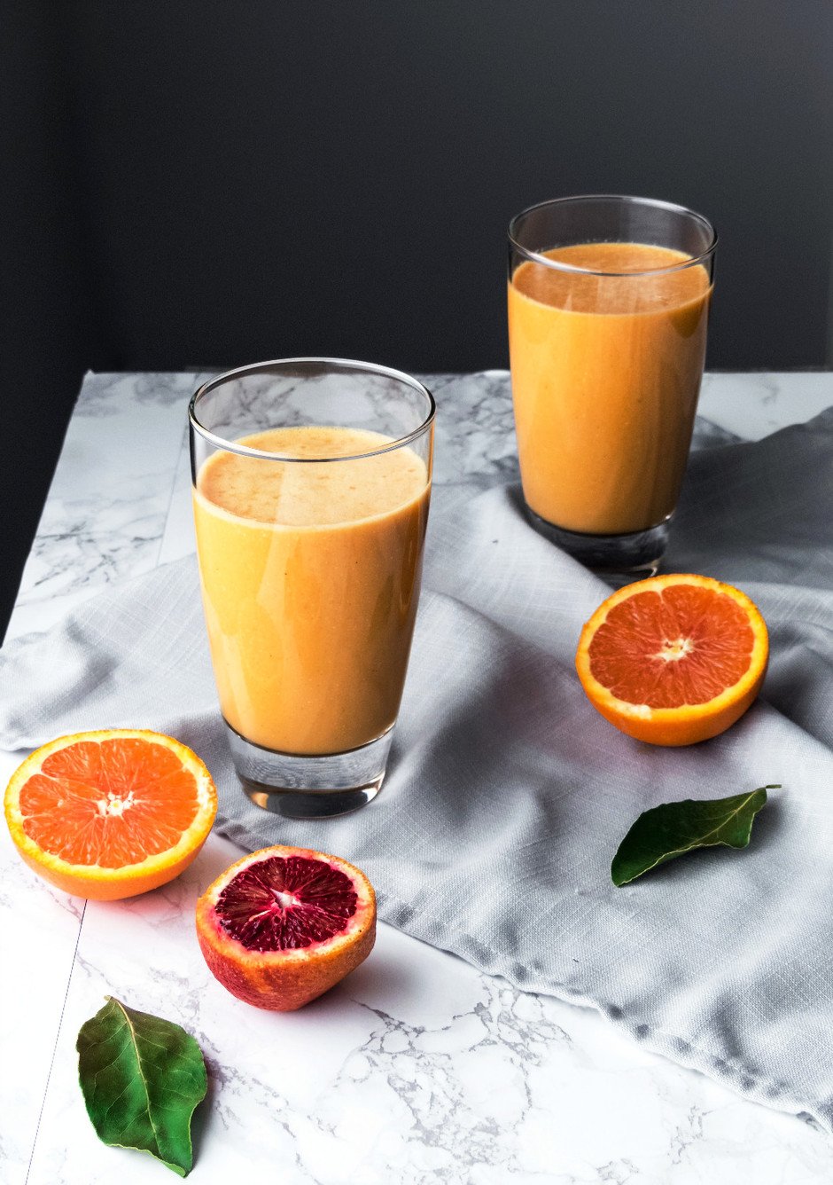 Angled overhead image of two glasses of citrus and root sunshine smoothie with oranges on the left and a blue linen.