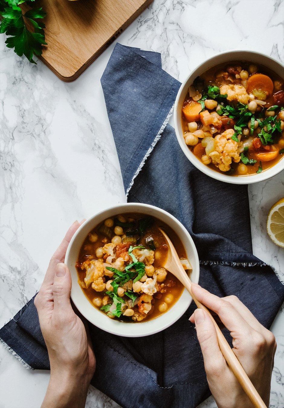 two hands holding a bowl and spoon of soup on a blue linen with another bowl on the right..