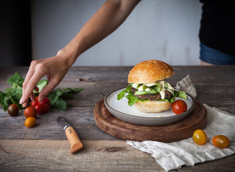 portrait of a veggie burger on a cutting board with a hand reaching for cherry tomatoes.