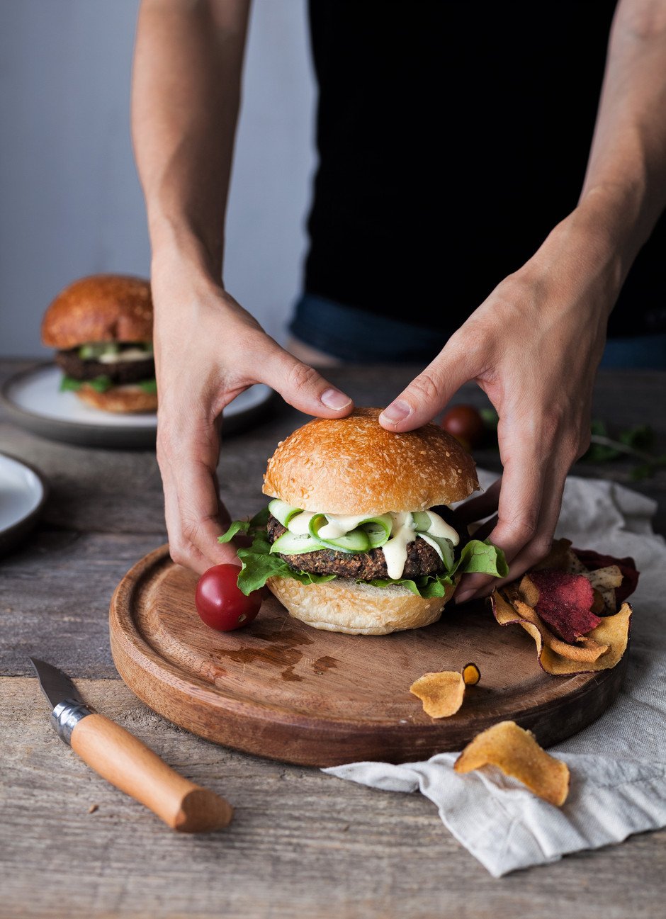 portrait of two hands holding a burger on a wooden board with chips and tomatoes.