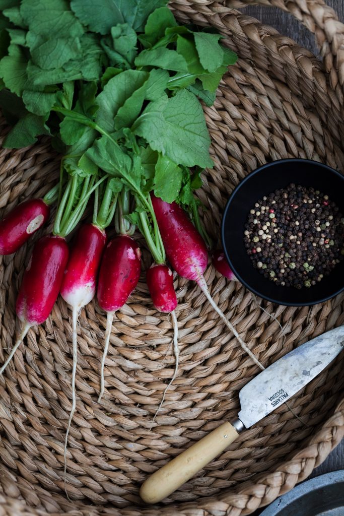 overhead of 6 radishes in a basket with a knife and peppercorns on the right side.