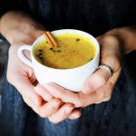 two hands cupping a mug of golden milk with a cinnamon stick.