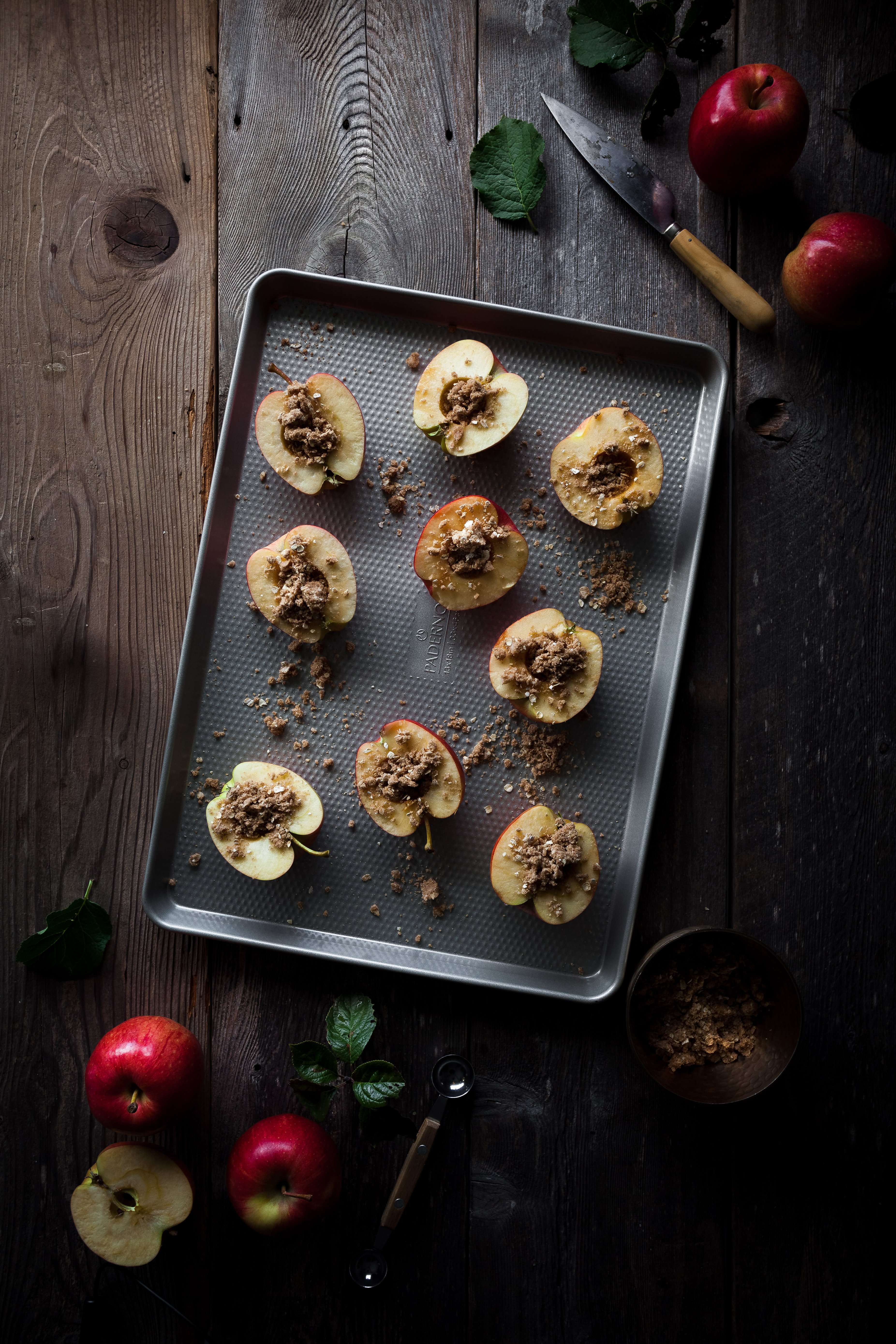 overhead image of apple halves filled with streusel on a baking sheet with whole apples and a knife arranged around it.