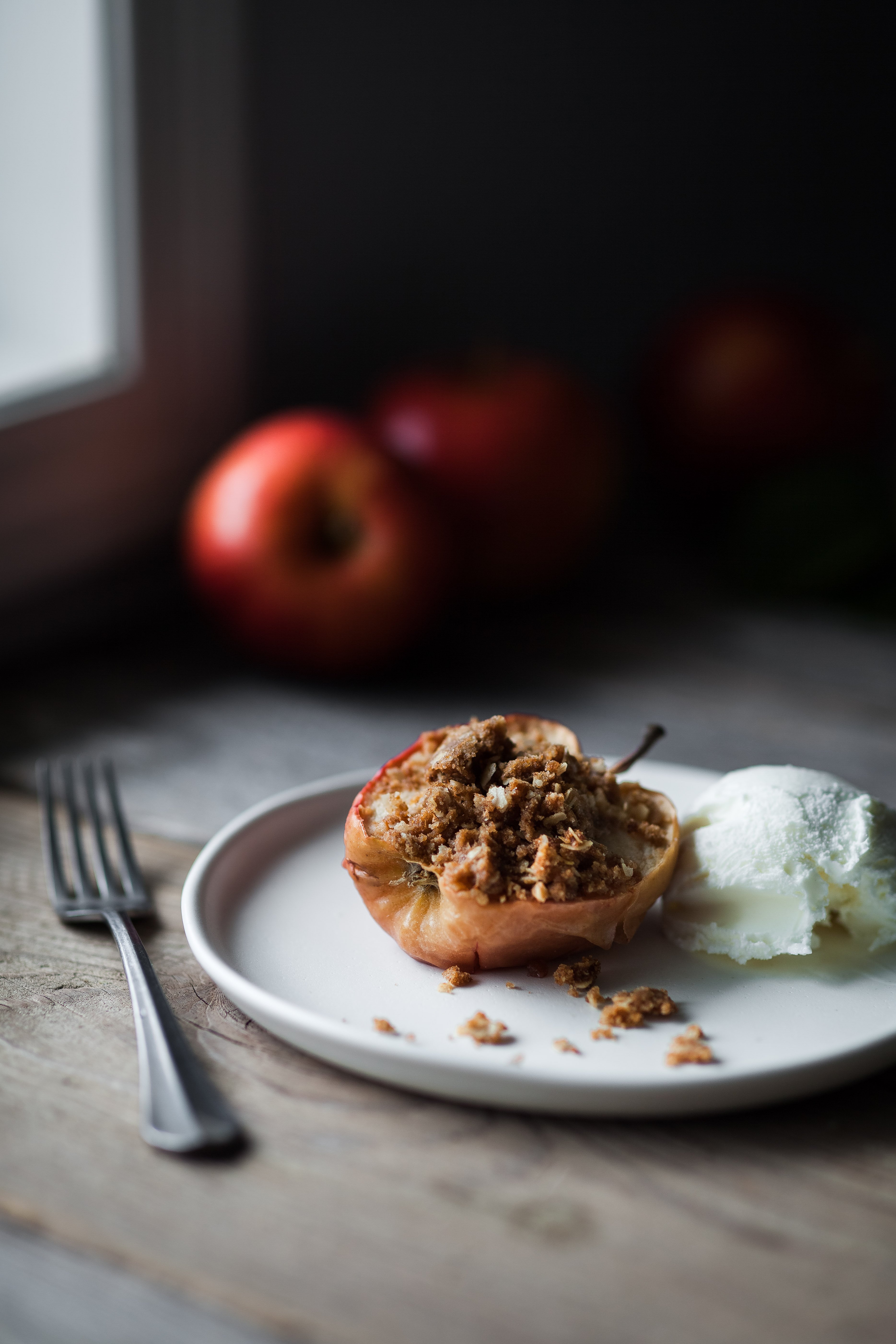 portrait of a Baked Apple with Oat Streusel on a white plate with ice cream with apples in the background.