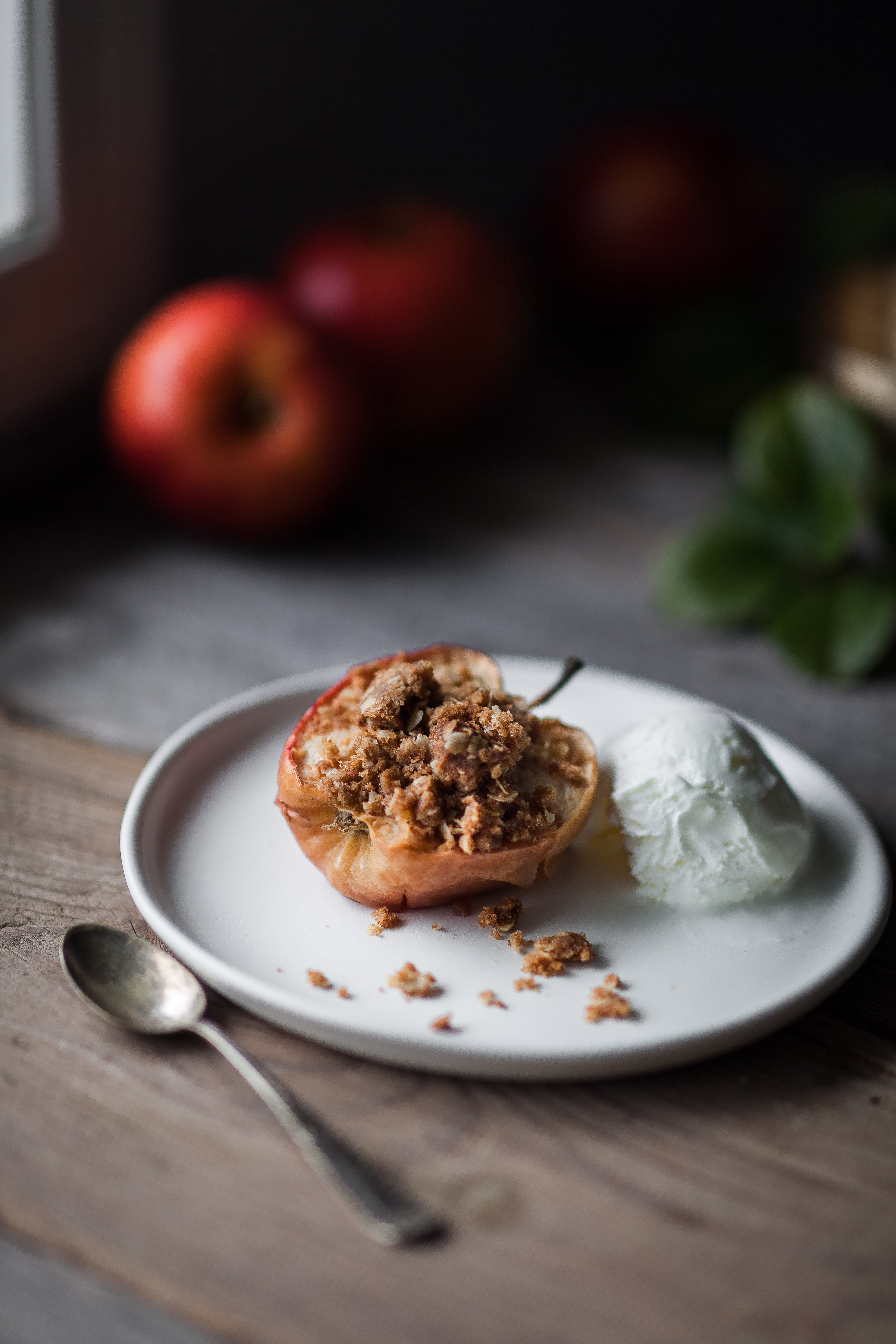 portrait of a Baked Apple with Oat Streusel on a white plate with ice cream with apples in the background.