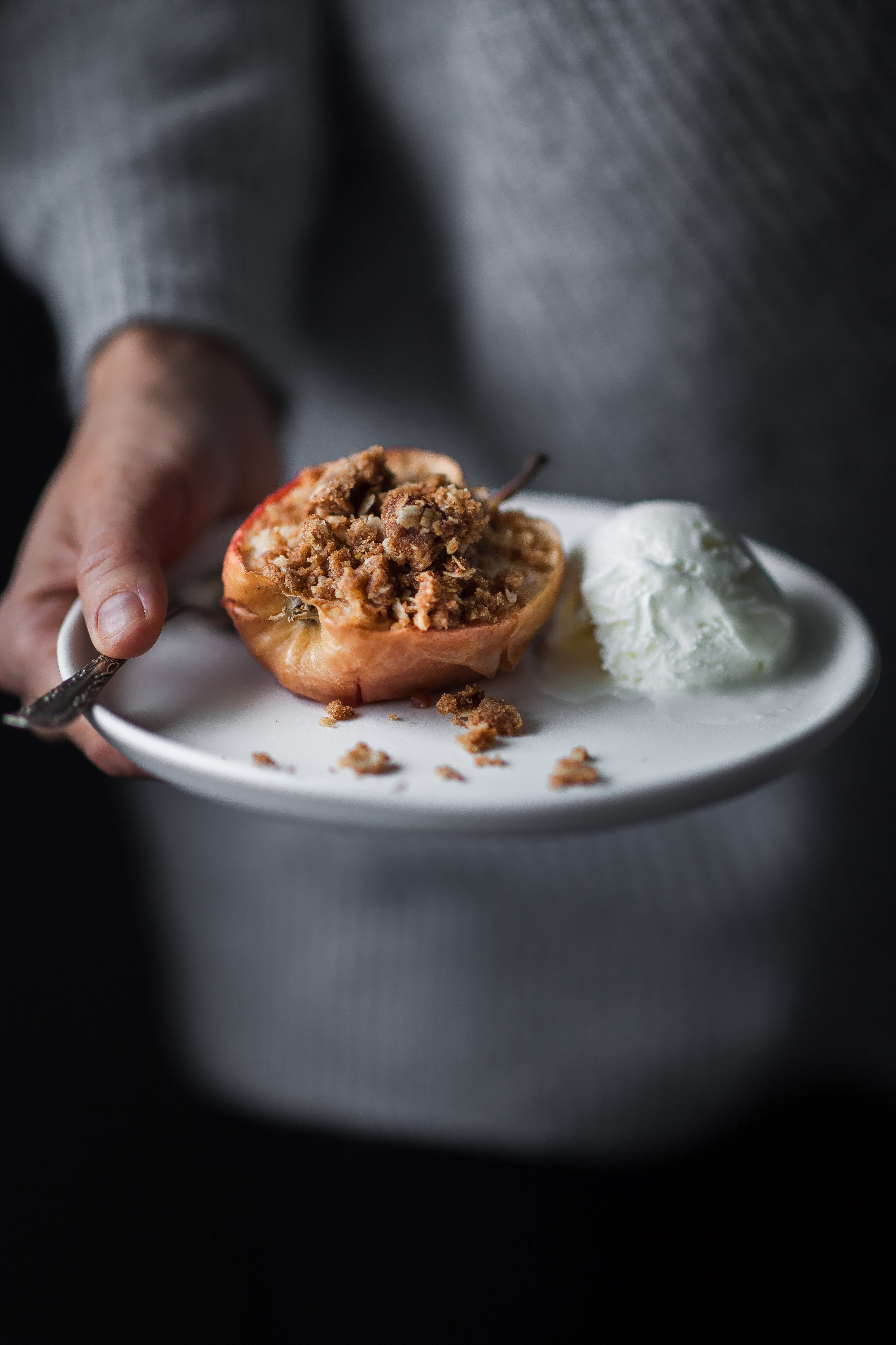 portrait of a person in a grey sweater holding a plate with a baked apple with ice cream.