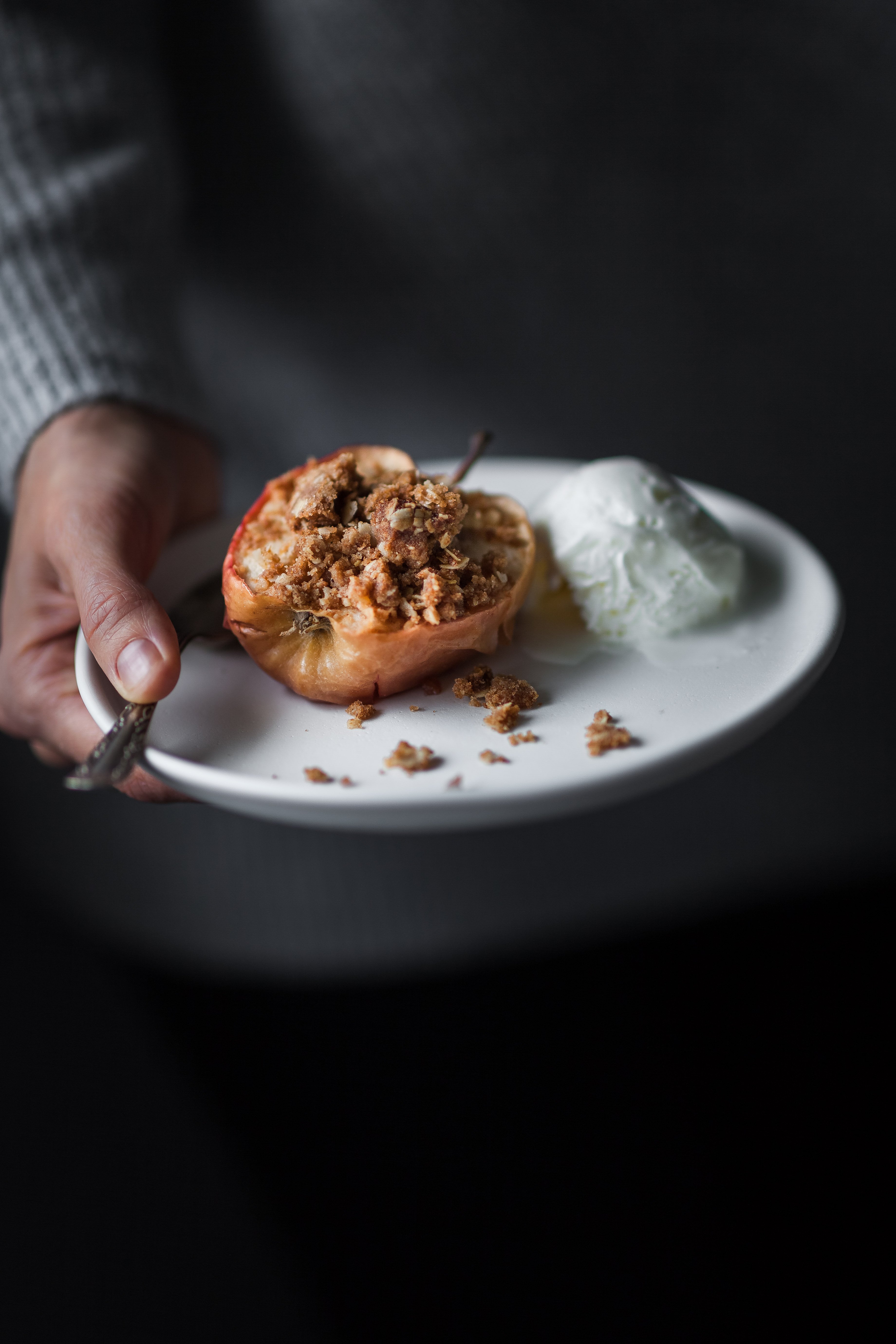 portrait of a person in a grey sweater holding a plate with a baked apple with ice cream.