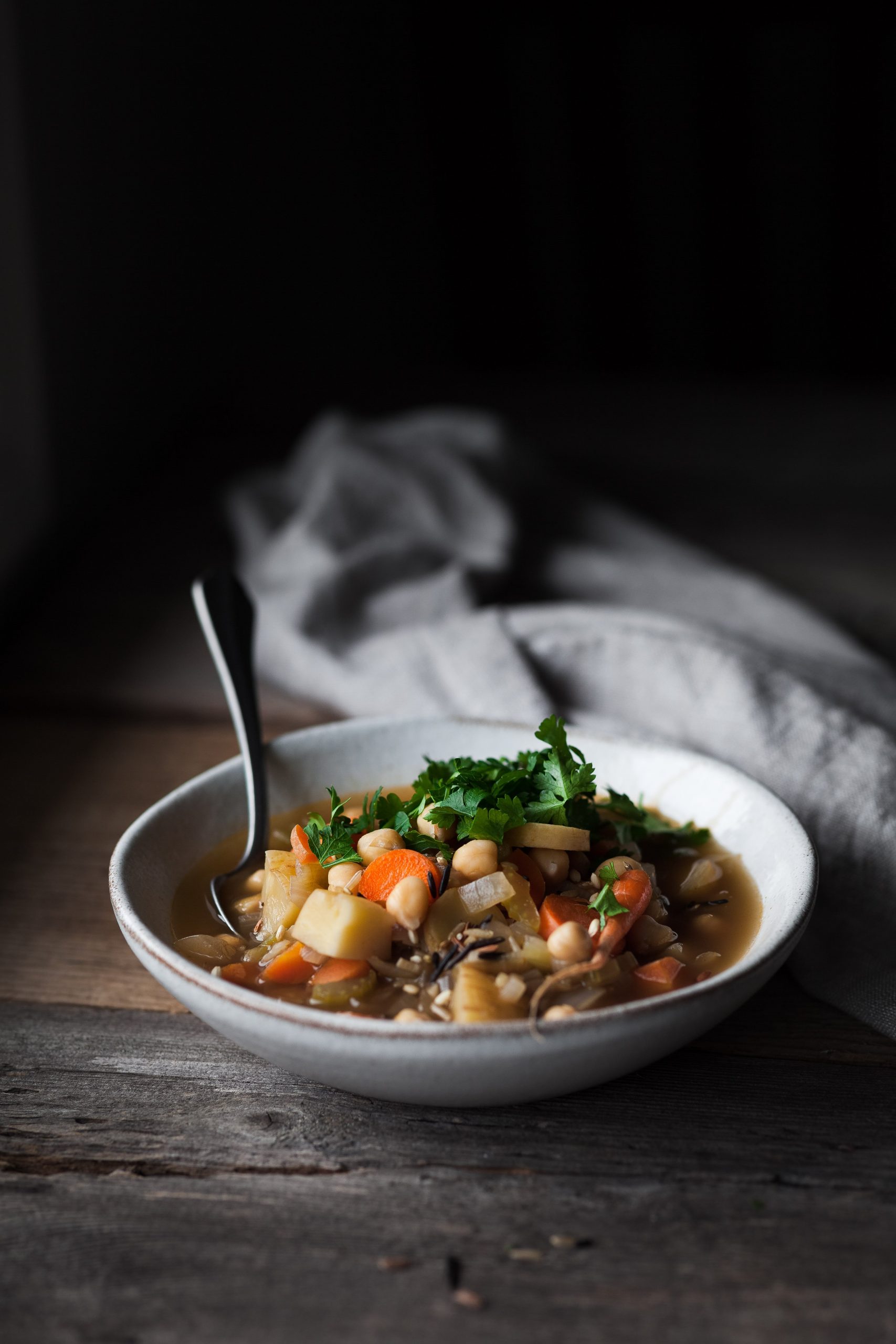 a head on image of a bowl of Winter Vegetable Soup with Chickpeas and parsley in a white ceramic bowl on a wood surface.