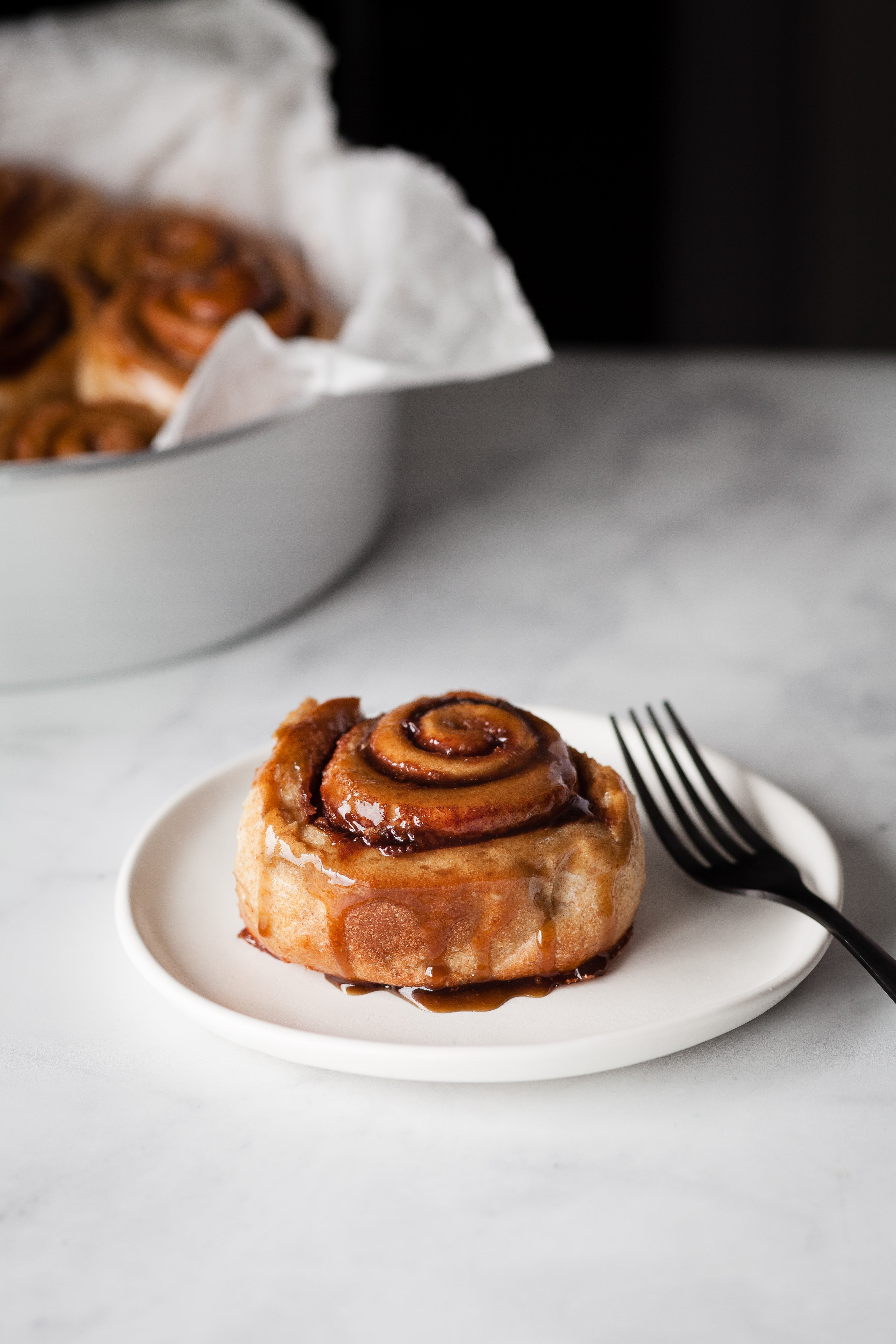 Head on shot of a vegan cinnamon roll on a white plate with a fork to the side.