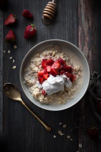 overhead view of bircher museli with strawberries and whipped cream on a wood surface with a gold spoon.
