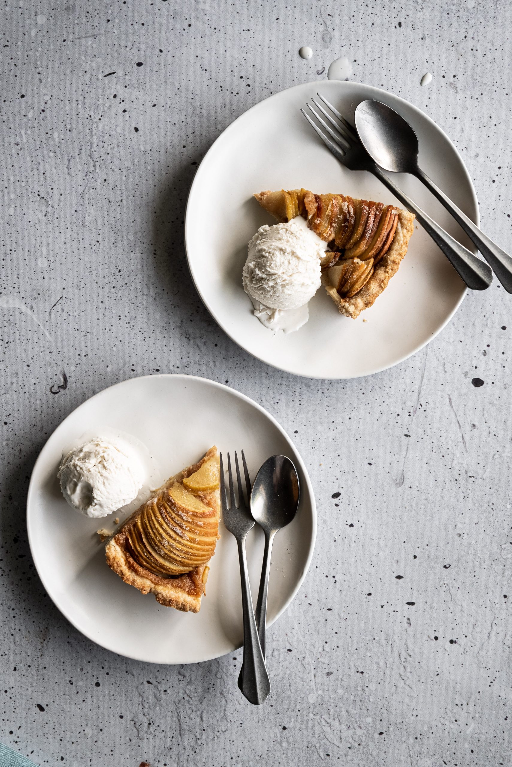 overhead image of two white plates with apple tart slices, ice cream and cutlery.
