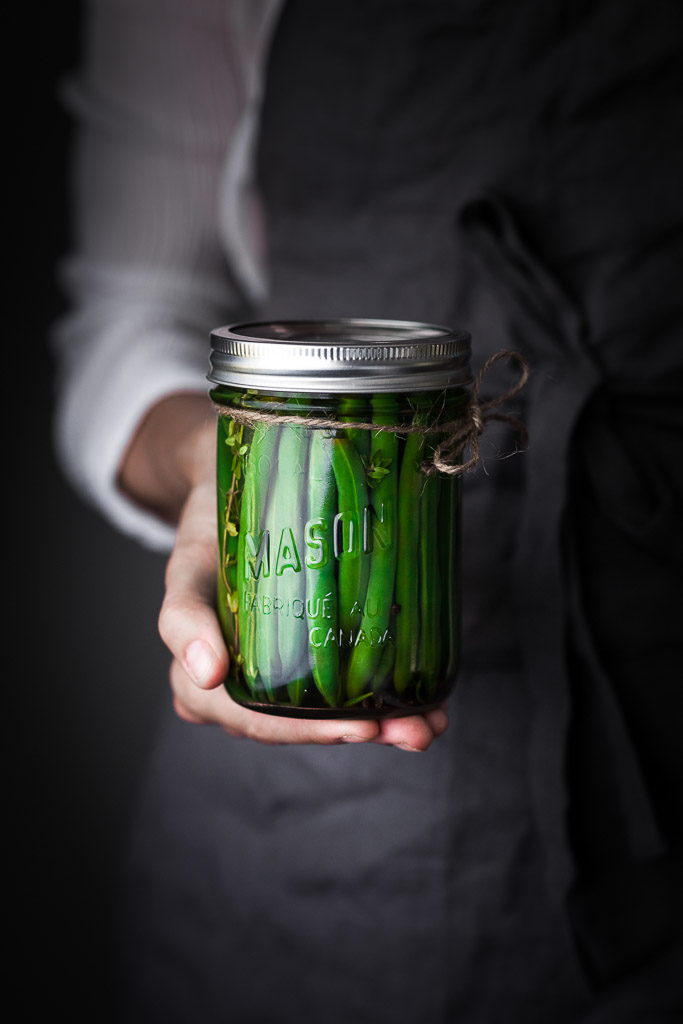 A person in a blue apron holding a jar of pickled green beans.