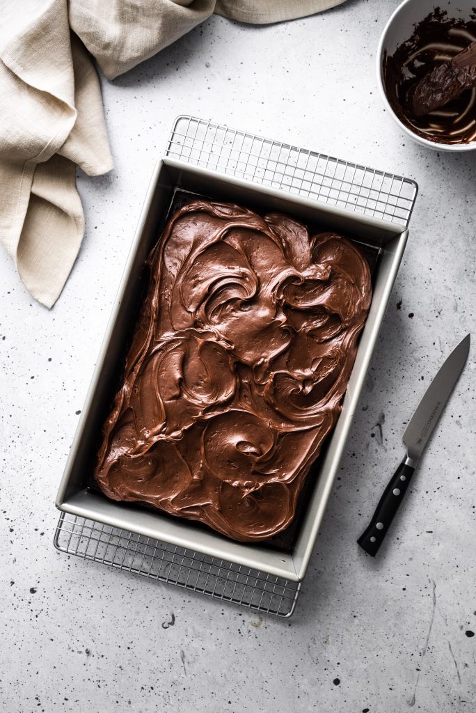 overhead image of a rectangular chocolate frosted cake on a grey surface with a beige linen and a knife.