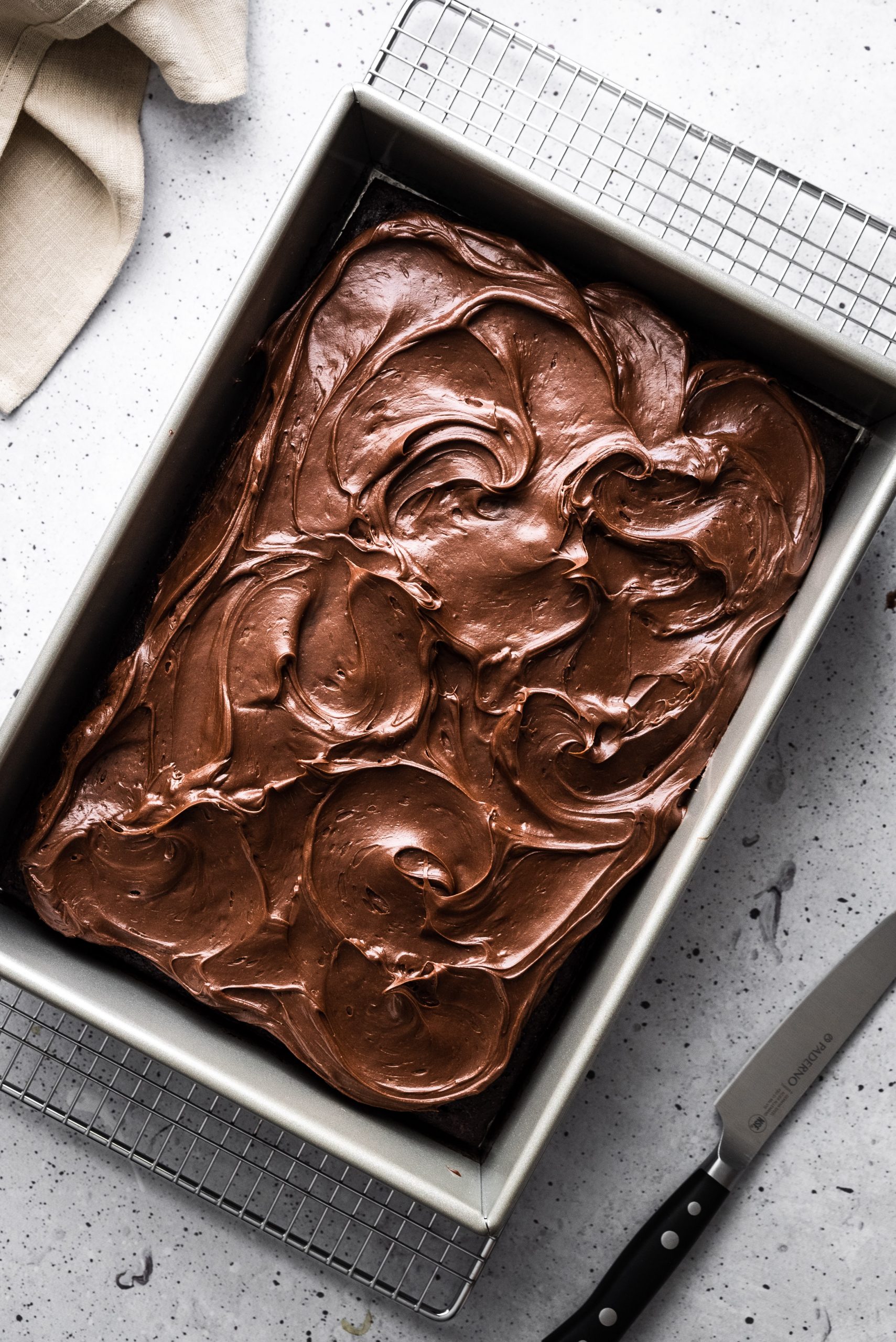 overhead image of a rectangular chocolate frosted cake on a grey surface with a beige linen and a knife.