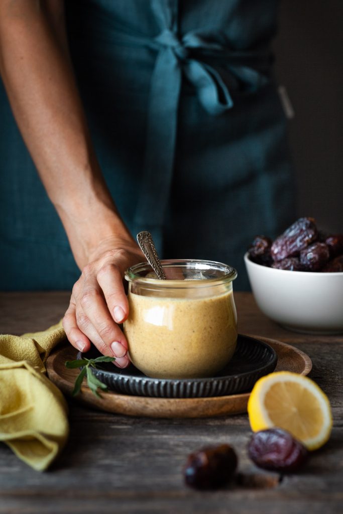 portrait view of a hand holding bowl of dressing with a yellow linen on the side and a bowl of dates in the background.