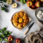 overhead view of an apple tarte tatin with a linen, apples and leaves and whipped cream on a marble surface.