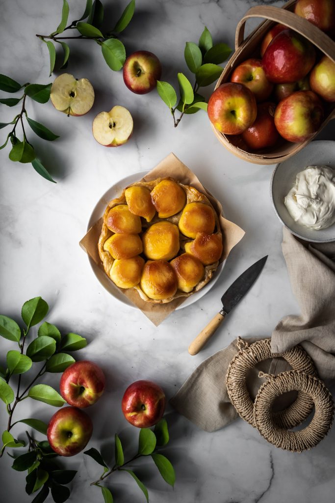 overhead view of an apple tarte tatin with a linen, apples and leaves and whipped cream on a marble surface.