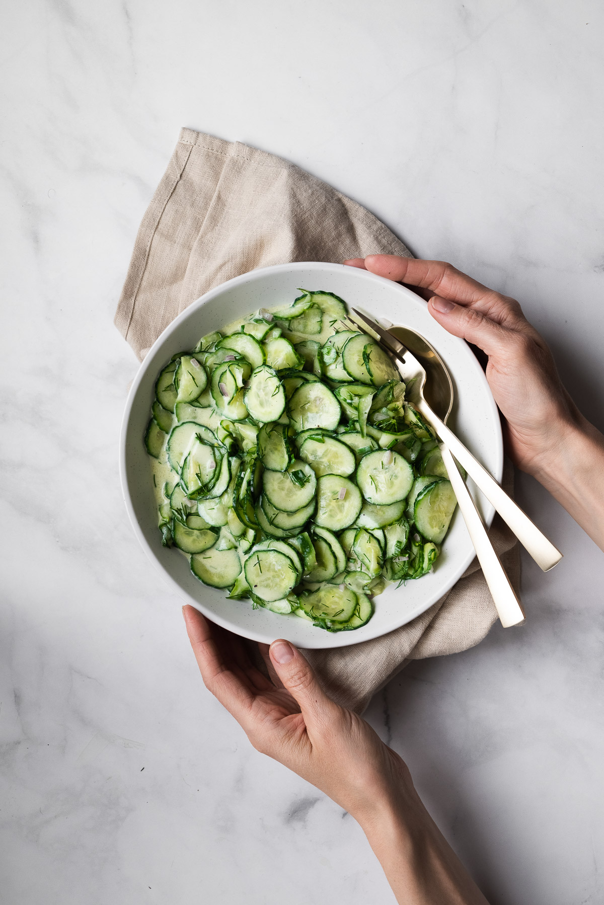 Overhead view of two hands holding a large bowl of vegan German cucumber salad.